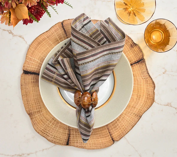 A Windsor Napkin from Kim Seybert in a multicolored striped linen is paired with a modern napkin ring and placed on a white plate with a gold rim, set atop a wooden slice charger. Nearby, two amber glasses and autumn flowers in neutral tones are arranged on a marble surface.