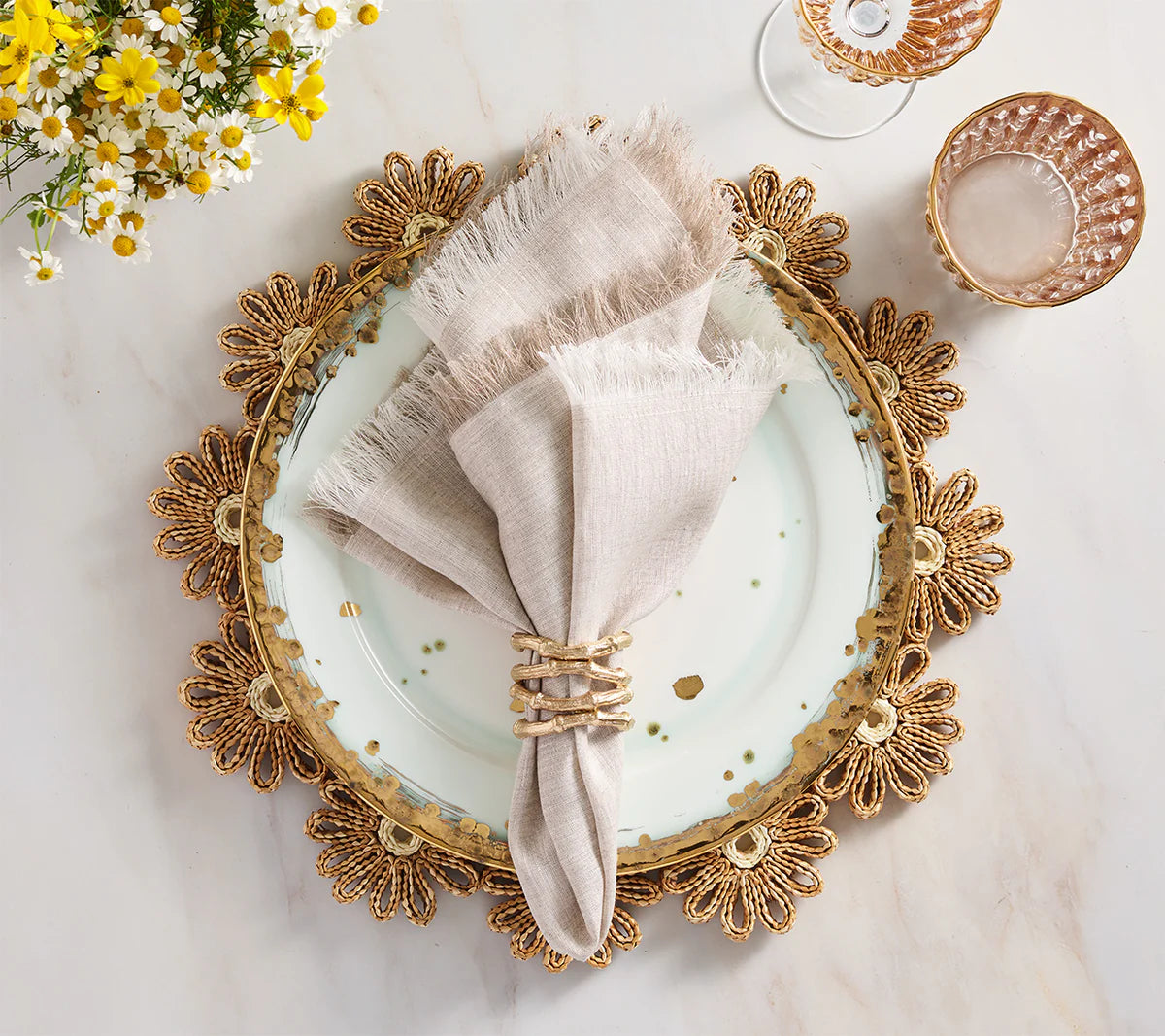 A white plate with gold accents sits on a floral gold placemat, creating charming farmhouse table decor. Resting on the plate is a Kim Seybert Chambray Fringe Napkin from the Set of 4, neatly placed in a woven ring. Nearby, crystal glasses and a yellow and white flower bouquet enhance the elegance of the marble table setting.