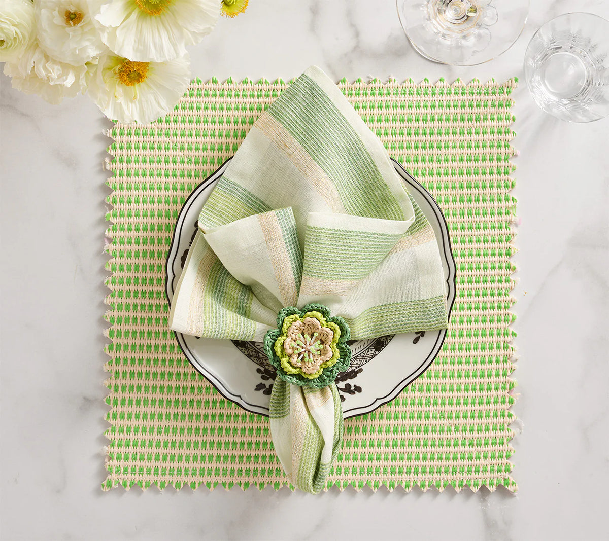 A green and white striped napkin with a floral ring rests on a decorative plate atop the Kim Seybert Duet Placemat Set of 4 - Blue & Green. White flowers and glassware add elegance to these sophisticated table settings on marble.