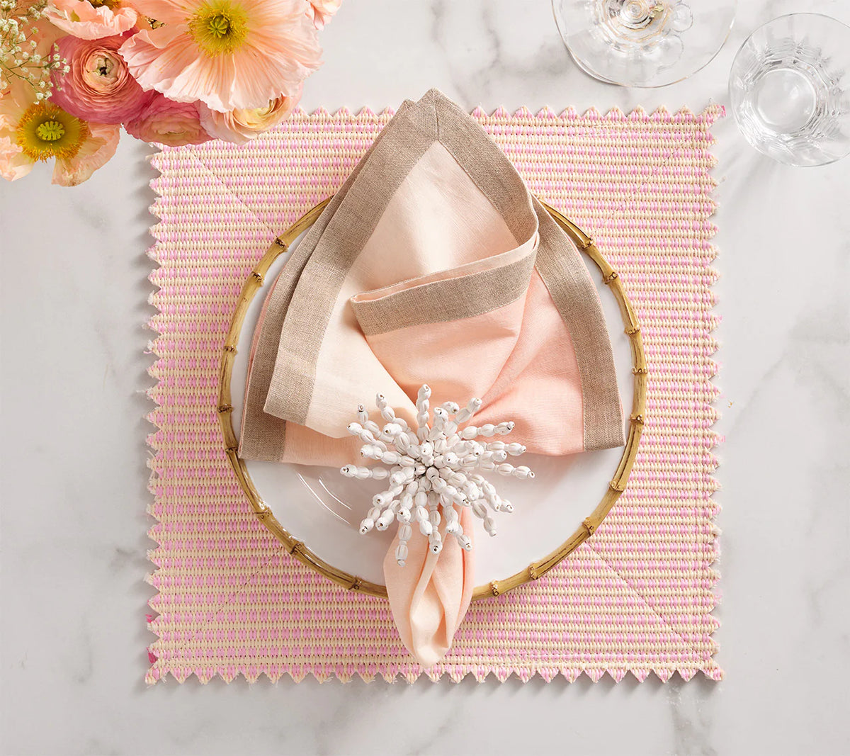 A table setting with a white plate on a bamboo-style charger, layered peach and tan napkins, a white beaded napkin ring, wine glasses, and peach flowers sits atop the Kim Seybert Duet Placemat Set of 4 - Blush & Pink on marble.