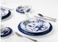 A place setting featuring Ginori 1735 Oriente Italiano Indaco plates with blue floral patterns, solid blue charger plates, a teacup and saucer, a glass, and silverware on a white tablecloth.