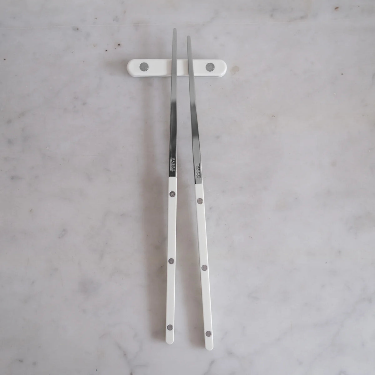 Two Bistrot Solid Chopsticks S/2 by Sabre, featuring white handles, rest on a matching white chopstick holder atop a light gray marble surface, showcasing elegant Asian tableware.