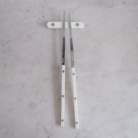 Two Bistrot Solid Chopsticks S/2 by Sabre, featuring white handles, rest on a matching white chopstick holder atop a light gray marble surface, showcasing elegant Asian tableware.