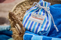 A blue and white striped drawstring pouch labeled "Southampton Beach Backgammon" by G. McGee sits on a plush cotton terry towel in a woven straw basket, evoking a summery beach vibe.