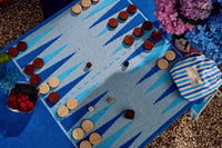 The G. McGee Southampton Beach Backgammon set is displayed on a plush cotton terry beach towel, beside a basket of mixed berries, a blue-and-white striped bag, and a bouquet of pink and blue flowers on pebbles.