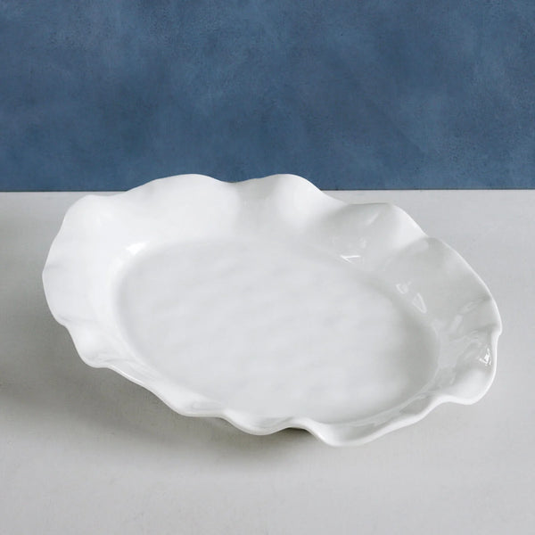 A white, oval Vida Havana Tray by Beatriz Ball with a wavy, scalloped edge rests on a light surface against a blue textured background, showcasing the sophisticated design of this luxury melamine platter.