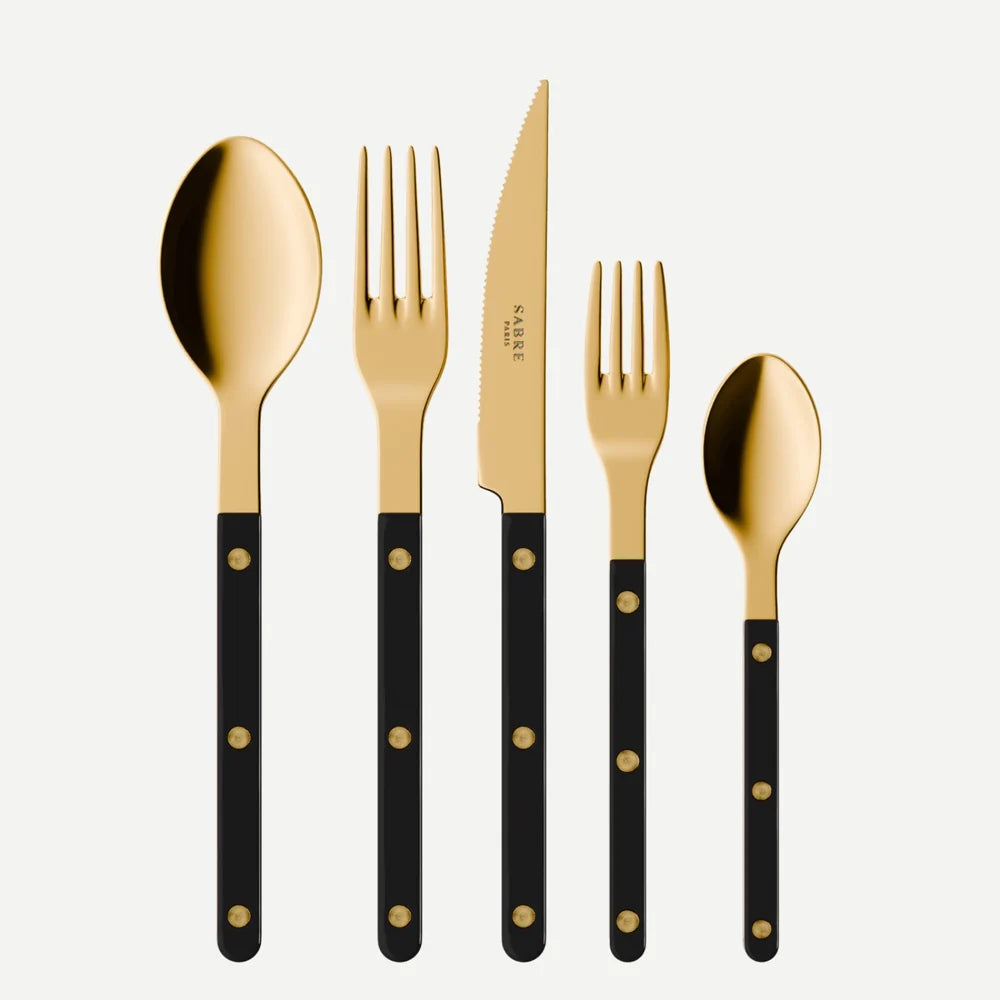 The Sabre Bistrot Gold 5-piece flatware set in Black features a large spoon, dinner fork, steak knife, salad fork, and small spoon, each with gold heads and black handles, arranged side by side on a white background.