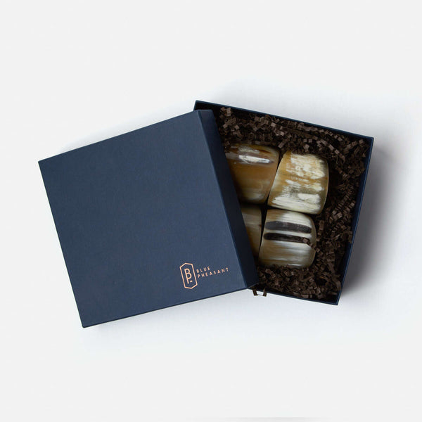 A navy blue Blue Pheasant box, partially open on a white background, holds four polished, handmade James Natural Horn Napkin Rings nestled in brown crinkle paper.