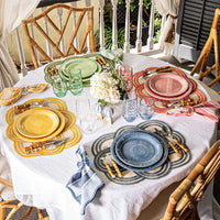 A round table set for four features colorful plates, matching glasses, striped napkins, and Juliska Scallop Stripe Placemats. A white tablecloth, center flowers, and bamboo chairs complete this bright outdoor scene with a touch of quatrefoil style.