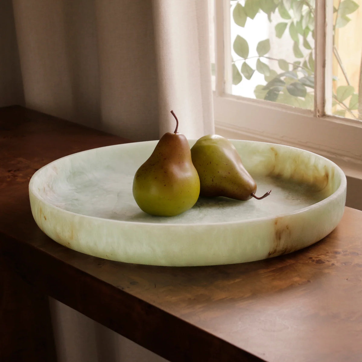 The Beatriz Ball Resin Giada Round Tray in Jade Swirl holds two pears on a wooden table by a window with sheer curtains, with leafy branches visible outside.