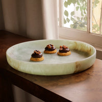 The Beatriz Ball Resin Giada Round Tray in Jade Swirl holds three cookies topped with chocolate and nuts on a wooden table by a window, where soft light streams through sheer curtains and leafy plants are visible outside.