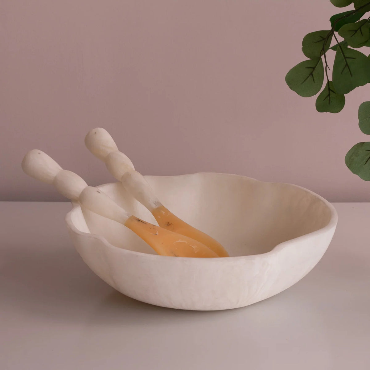 The Beatriz Ball Resin Sorbet Salad Server Set - Coconut, featuring a white scalloped-edge bowl and two matching servers, is displayed on a light surface with green leaves overhead against a pale mauve wall.
