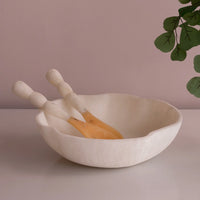 The Beatriz Ball Resin Sorbet Salad Server Set - Coconut, featuring a white scalloped-edge bowl and two matching servers, is displayed on a light surface with green leaves overhead against a pale mauve wall.