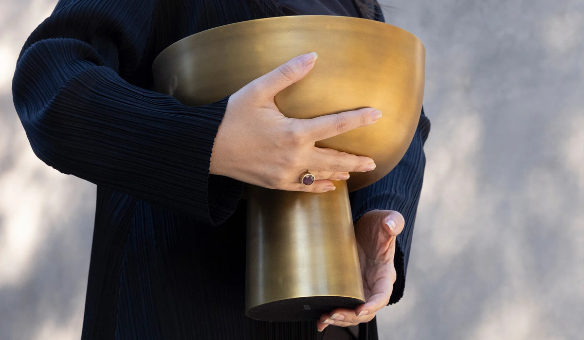 A person wearing a dark, textured outfit holds the large, handcrafted Kara Brass Footed Bowl by Dumae with both hands. A ring with a dark stone adorns one finger. The neutral, blurred background draws attention to this artisan home decor piece.