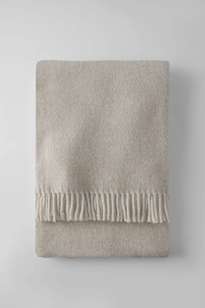 The A Soft Idea Tweed Weave Throw, in a neatly folded beige style with a fringe on one edge, is displayed on a light gray surface.
