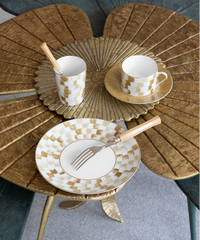 The Marie Daâge Zephyr 1 Dinnerware set by Marie Daâge features a gold and white checkered hand-painted porcelain plate, cup, and saucer, elegantly accompanied by wooden-handled cutlery on a gold flower-shaped table. Made in France, this sophisticated ensemble adds refined charm to any dining setting.