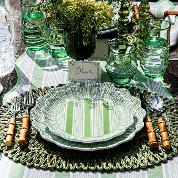 A table setting features Juliska’s Cabana Stripe Melamine Dinnerware Set of 4 - Seagrass, paired with bamboo-handled cutlery, green glassware, flowers, and an “Olivia” place card. These shatterproof dinner plates are dishwasher safe.