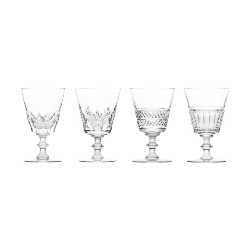 A set of four Queen’s Hall glasses by Saint-Louis/Hermès, each featuring a distinct etched or cut pattern on the bowl and a short, rounded pedestal base, displayed in a row against a white background—capturing the timeless elegance of tableware once found in the Palace of Versailles.