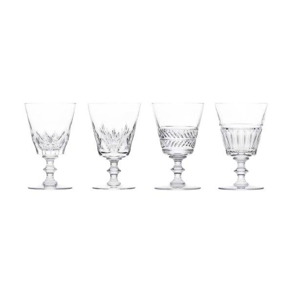 A set of four Queen’s Hall glasses by Saint-Louis/Hermès, each featuring a distinct etched or cut pattern on the bowl and a short, rounded pedestal base, displayed in a row against a white background—capturing the timeless elegance of tableware once found in the Palace of Versailles.