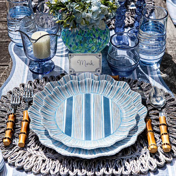 A table setting with Juliska Cabana Stripe Melamine Dinnerware Set of 4 - Ocean, bamboo cutlery, blue glassware, a candle in a blue holder, floral centerpiece, Mark's place card on a woven placemat and striped tablecloth.