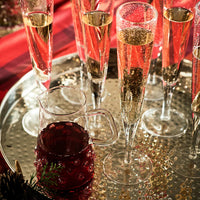 A tray of tall champagne flutes with sparkling wine and an Abigails Swirl Glass Pitcher filled with a dark red drink, arranged on a festive red tablecloth.