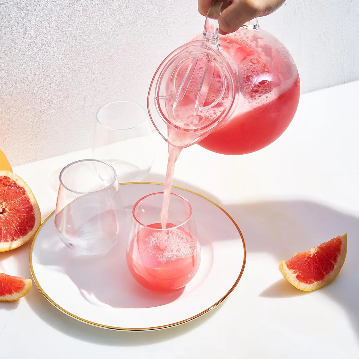 A hand pours pink juice from The Wine Savant/Clingbrands Unbreakable Brighton Acrylic Clear Lidded Pitcher into a glass on a white plate with a gold rim, surrounded by empty glasses and grapefruit wedges on a white surface.