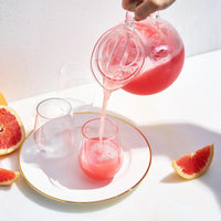 A hand pours pink juice from The Wine Savant/Clingbrands Unbreakable Brighton Acrylic Clear Lidded Pitcher into a glass on a white plate with a gold rim, surrounded by empty glasses and grapefruit wedges on a white surface.
