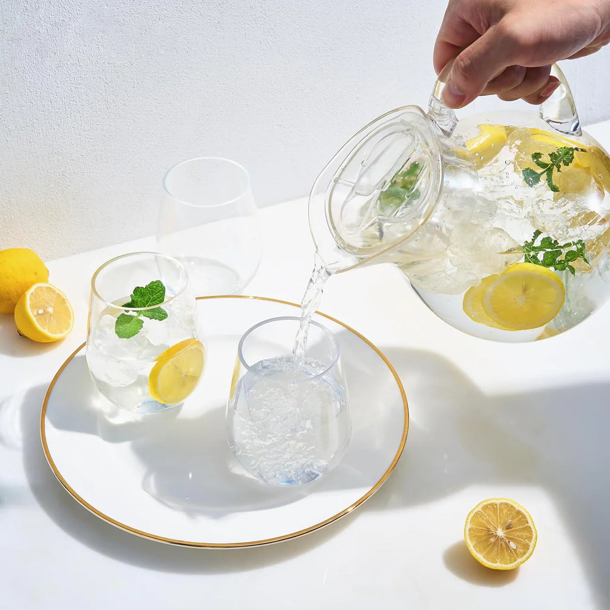 A hand pours water from The Wine Savant/Clingbrands Unbreakable Brighton Acrylic Clear Lidded Pitcher with lemon slices and mint into a glass on a white plate. Nearby are two more glasses with lemon, mint, and ice, plus lemons on a white surface.