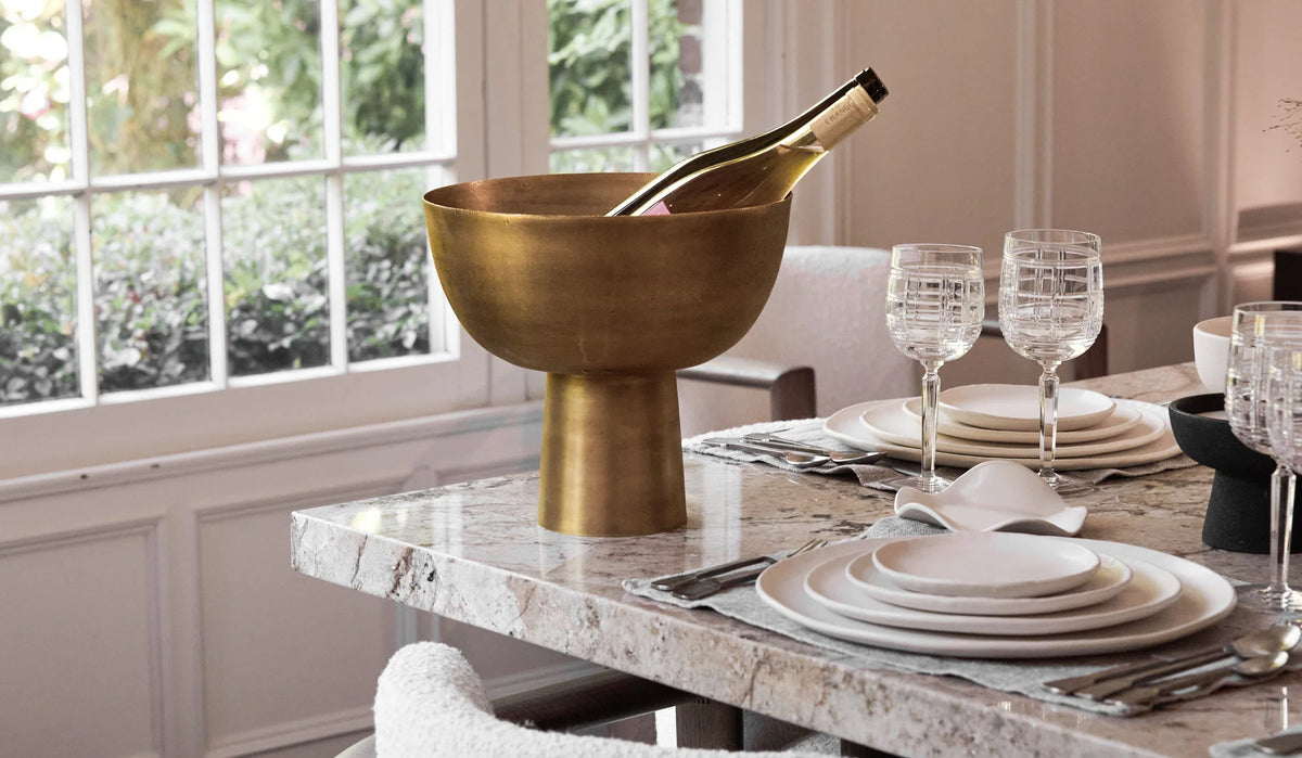 A Dumae Kara Brass Footed Bowl holding a bottle stands on a marble dining table set with white dishes, glasses, and silverware near a window overlooking greenery, creating an inviting scene of artisan home decor.