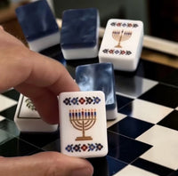 A hand holds a white acrylic Mahjong tile from the Brilliant Nights Hanukkah in Gift Box set, featuring a gold menorah with blue and purple candles, surrounded by other tiles on a checkered board.