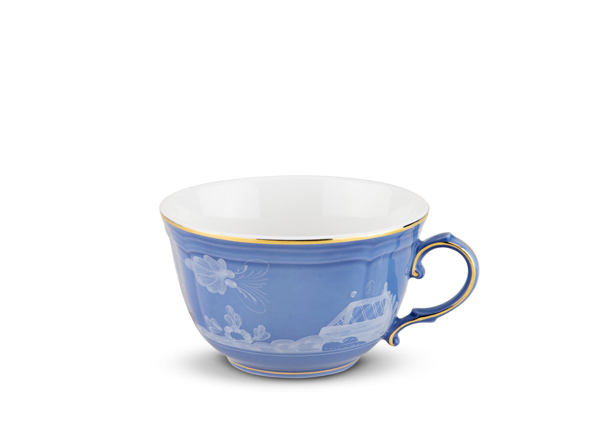 The Oriente Italiano Pervinca teacup by Ginori 1735 is a blue porcelain cup with a white and 24-karat gold rim, featuring a white scenic bridge, clouds, and flower design, styled like the Oriente Italiano line. Shown on a plain white background.