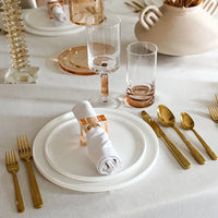 A chic table setting featuring white plates, a rolled napkin held by the Lucite Cube Napkin Ring Set of 4 - Amber from BT- Shalom, gold cutlery, clear and pink glasses, all displayed on a white tablecloth with stylish decorative accents in the background—an ideal showcase of contemporary table decor.