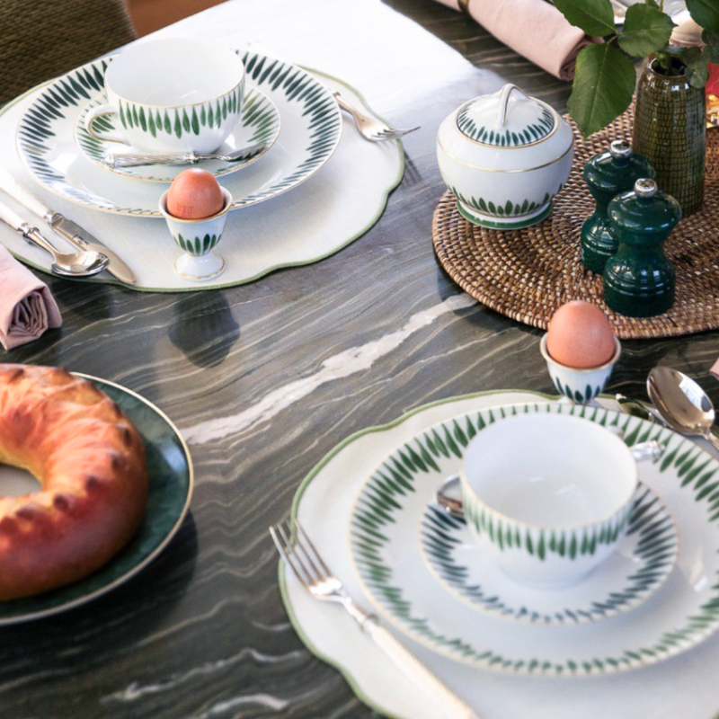 A table set with Marie Daâge Allee de Cypres 5 white dishes by Marie Daâge, green leaf patterns, bread, two eggs in egg cups, green glassware on a woven mat—all arranged on a dark marble tabletop.