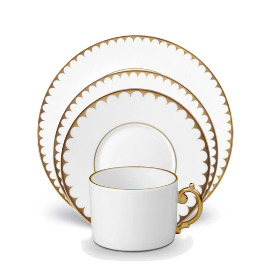 The L'Objet Aegean Filet Gold Dinnerware set includes a porcelain teacup, saucer, and two plates, each featuring scalloped 24k gold rims. The pieces are elegantly stacked with the cup in front of the plates.