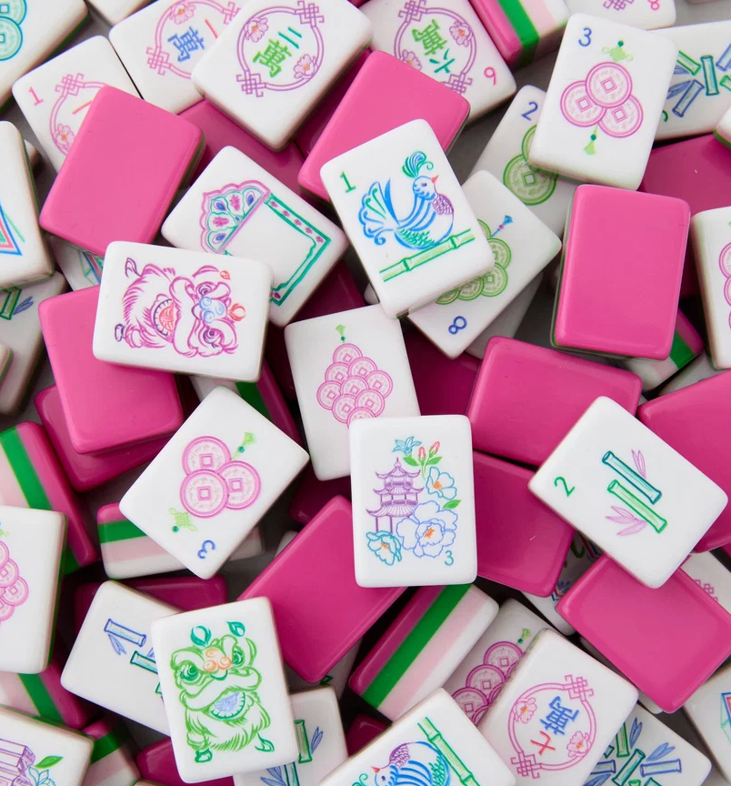 A scatter of vibrant Mahjong tiles from the Oh My Mahjong “Mahjong Tiles Set - Amara,” predominantly pink and white, features an array of Chinese characters, symbols, bamboo, birds, and flowers. The acrylic tiles are arranged haphazardly, with some facing up and others upside down.