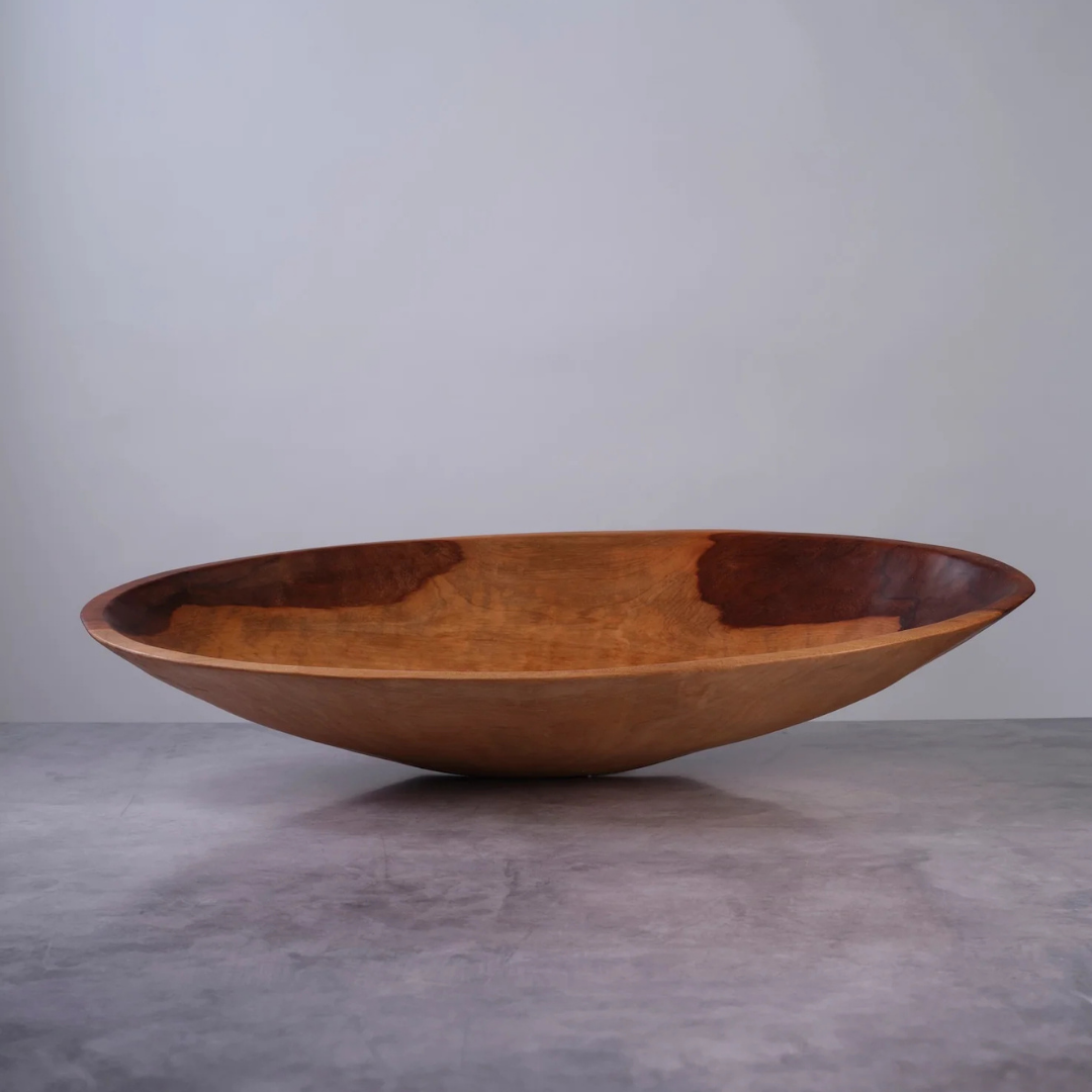 The Sobremesa Artisan Oval Wood Bowl - Medium 22″ is a large, shallow bowl hand-carved from sustainably sourced wood, featuring rich natural grain patterns and a food-safe finish. It sits on a gray surface against a plain light gray background.