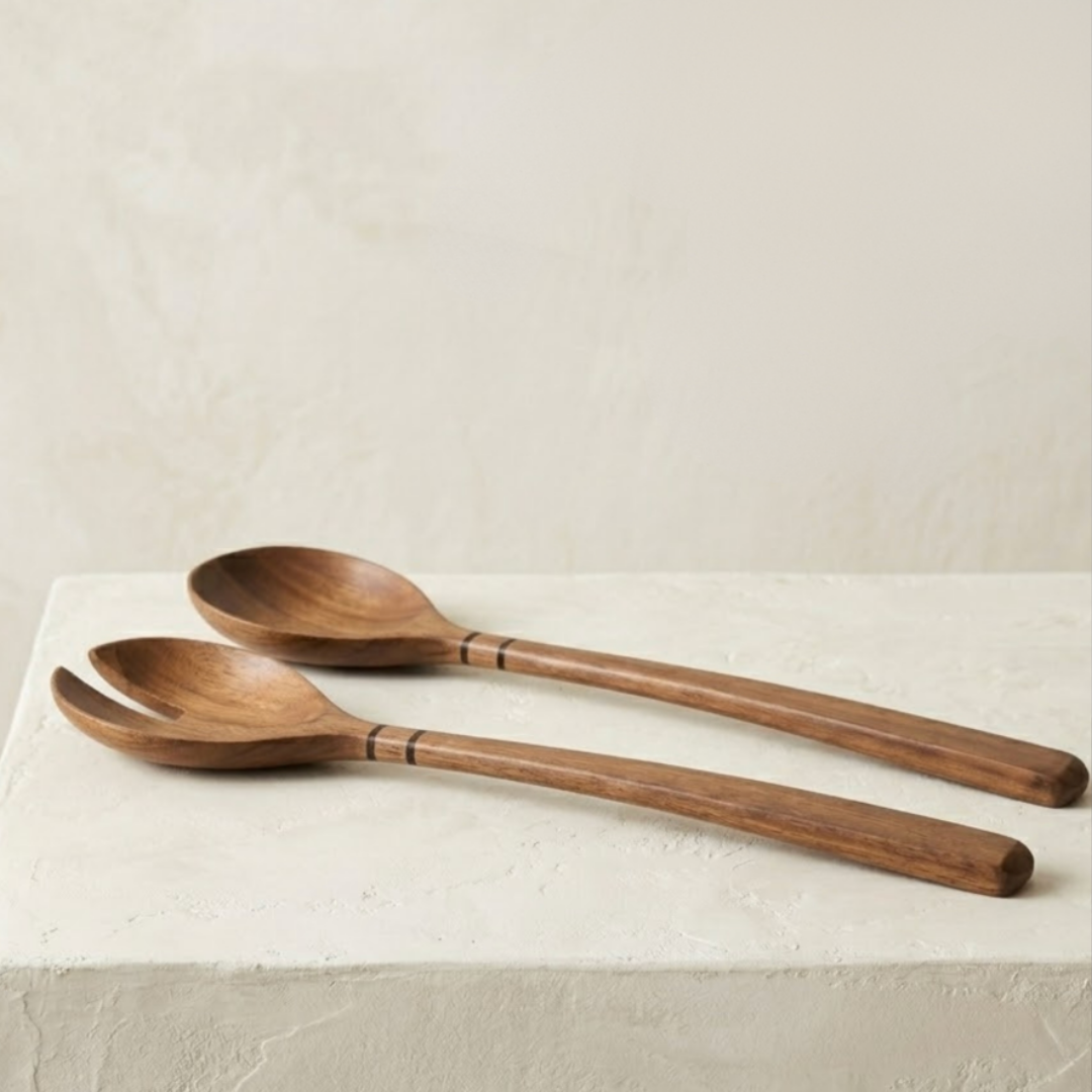 The Volare Serving Set by Be Home features two acacia wood salad servers—a spoon and a fork—with long handles adorned with decorative bands and a natural finish, displayed on a textured white surface against a light background.
