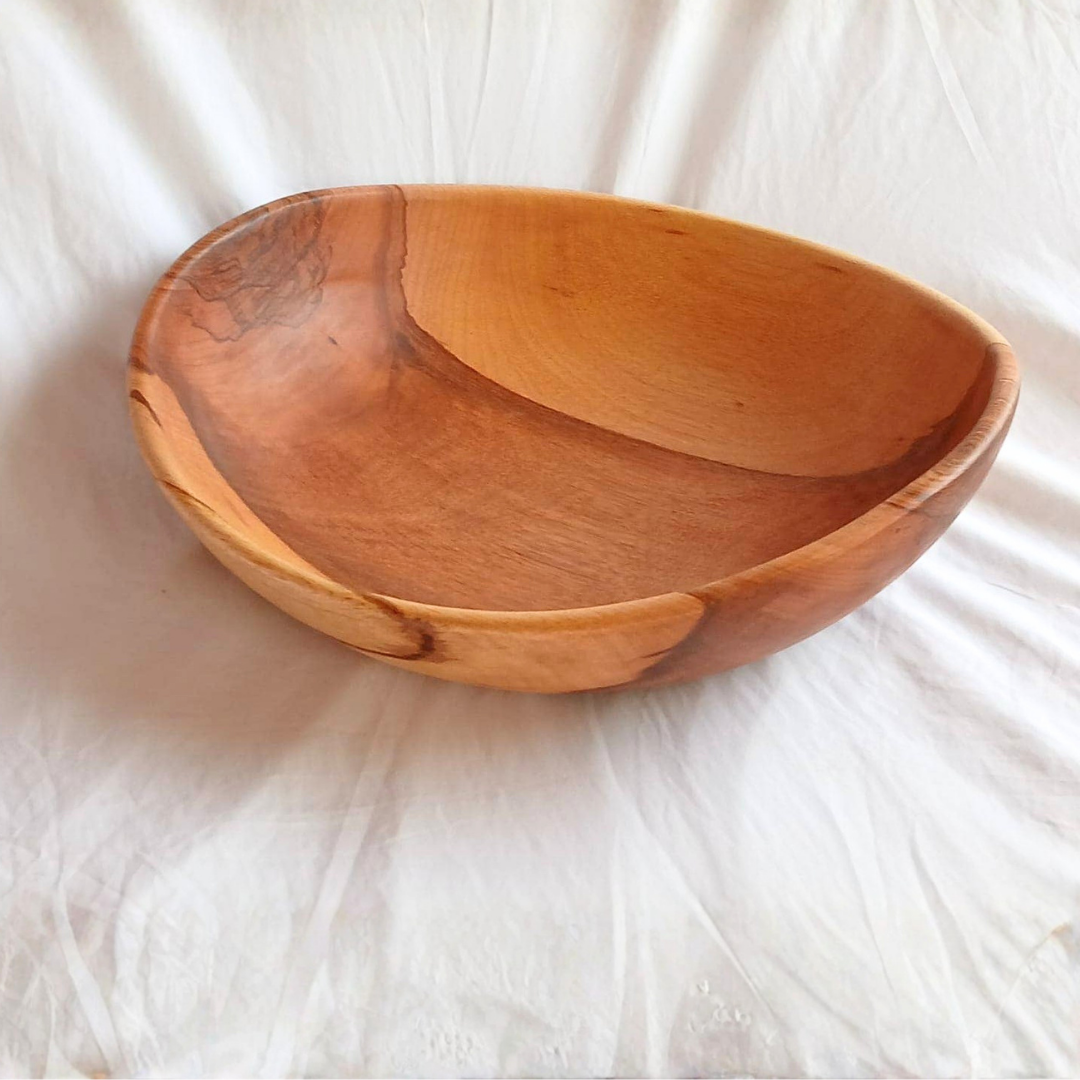 A large, shallow Sobremesa Artisan Hand Carved Wave Rim Wood Bowl - 17″ with a smooth, natural finish sits on a white fabric surface. Showcasing visible wood grain and an irregular, slightly oval shape, this handcrafted hardwood bowl exemplifies eco-friendly craftsmanship.