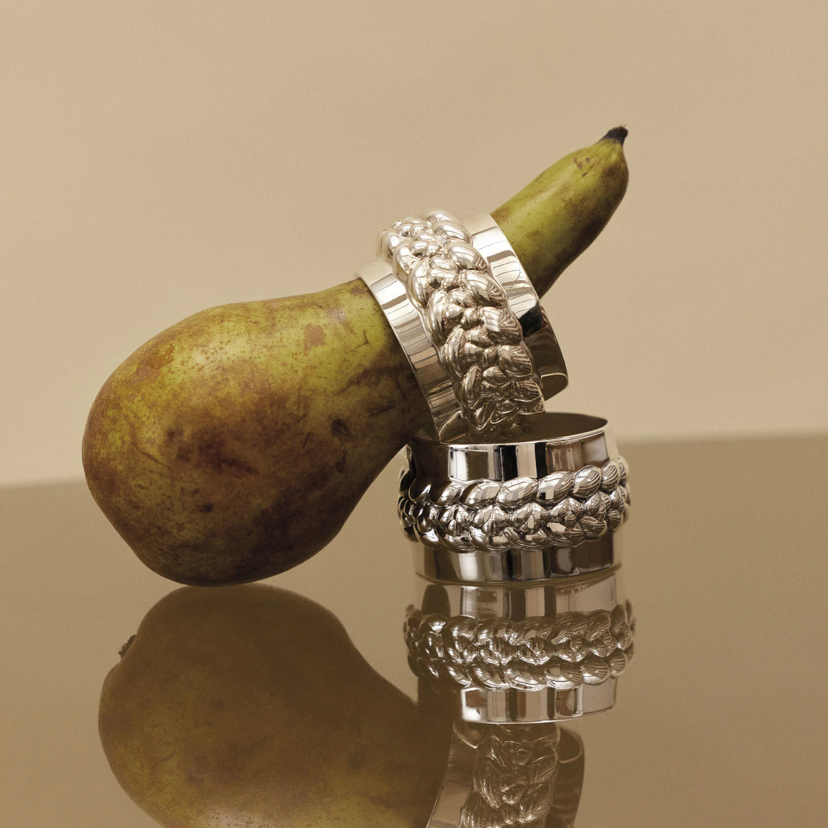 Two Christofle Babylone Silver Plated Napkin Rings with a braided texture, one encircling a pear and one resting below it on a reflective surface.
