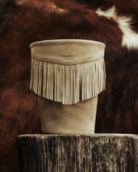 A mineral taupe leather Calamity Jane Candle - The Plains holder with fringe detail, resting on a tree stump.