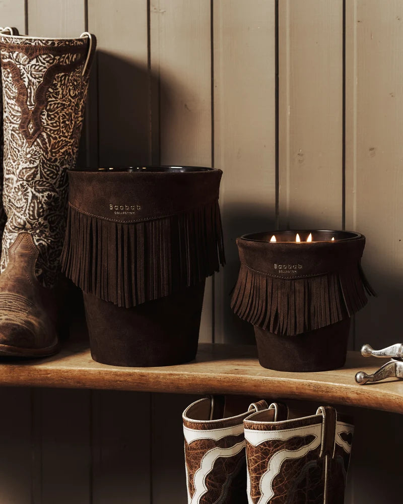 Two dark brown, fringed Calamity Jane Candle - Deadwood vessels sit on a wooden shelf next to a pair of cowboy boots.