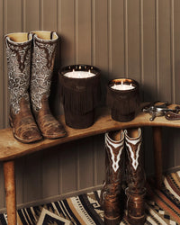 Two lit Calamity Jane Candle - Deadwood candles in fringed holders sit on a wooden bench beside two pairs of western-style cowboy boots.