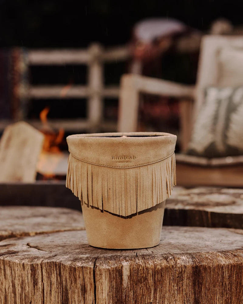 Calamity Jane Candle - The Plains mineral taupe leather candle holder with fringe detail resting on a tree stump.