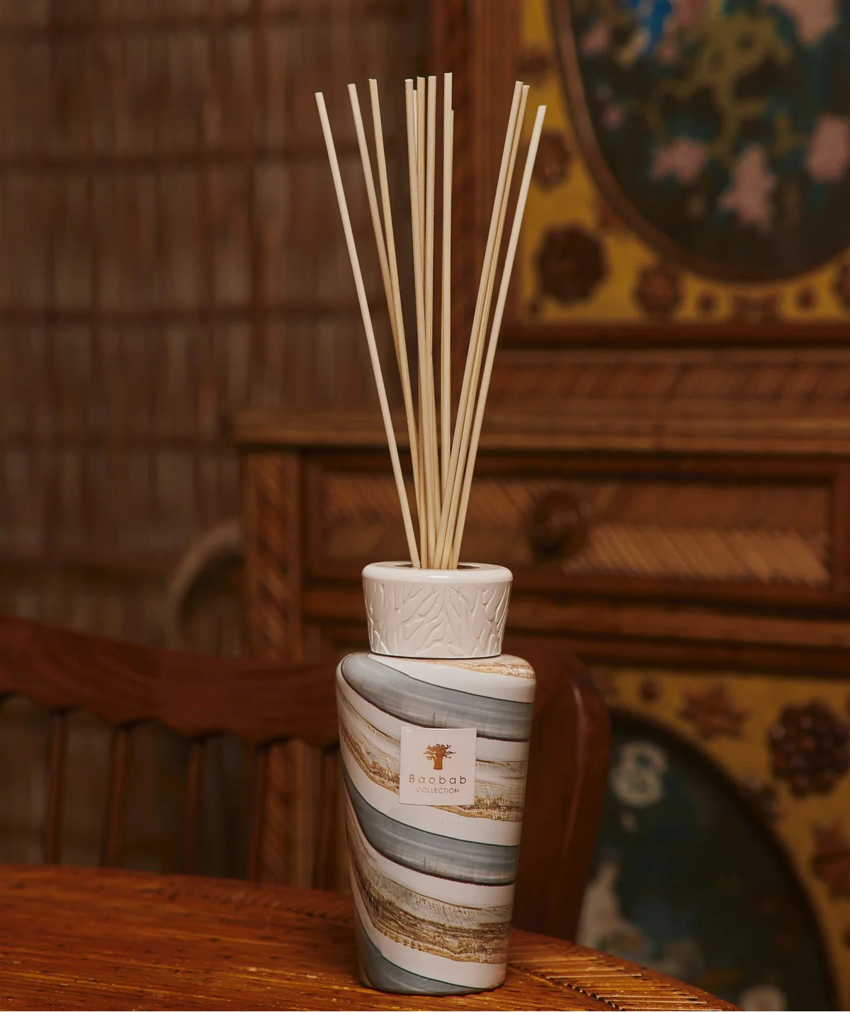 A Baobab Sand Sonora Diffuser, crafted as a ceramic reed diffuser complete with multiple wooden sticks, sits elegantly on a wooden table. In the background, a wooden chair and an intricately designed decorative cabinet add warmth and charm to your interior décor.