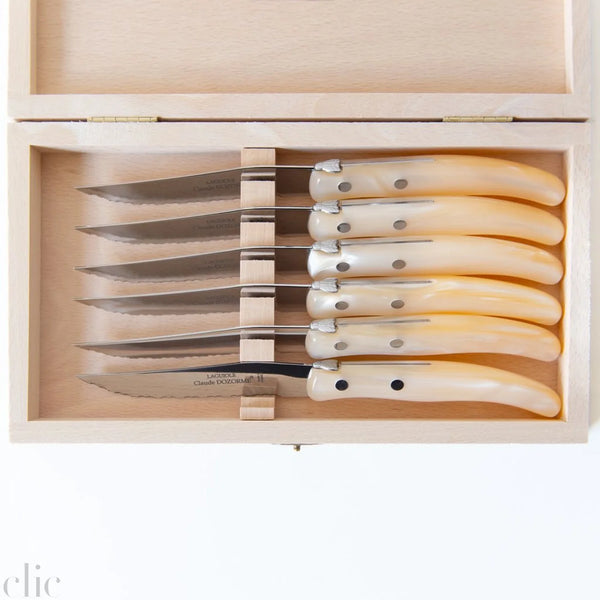 A wooden box containing a Claude Dozorme Berlingot Steak Knives s/6 Assorted set, featuring six French steak knives with polished, assorted handles and stainless steel blades, neatly arranged in individual slots.