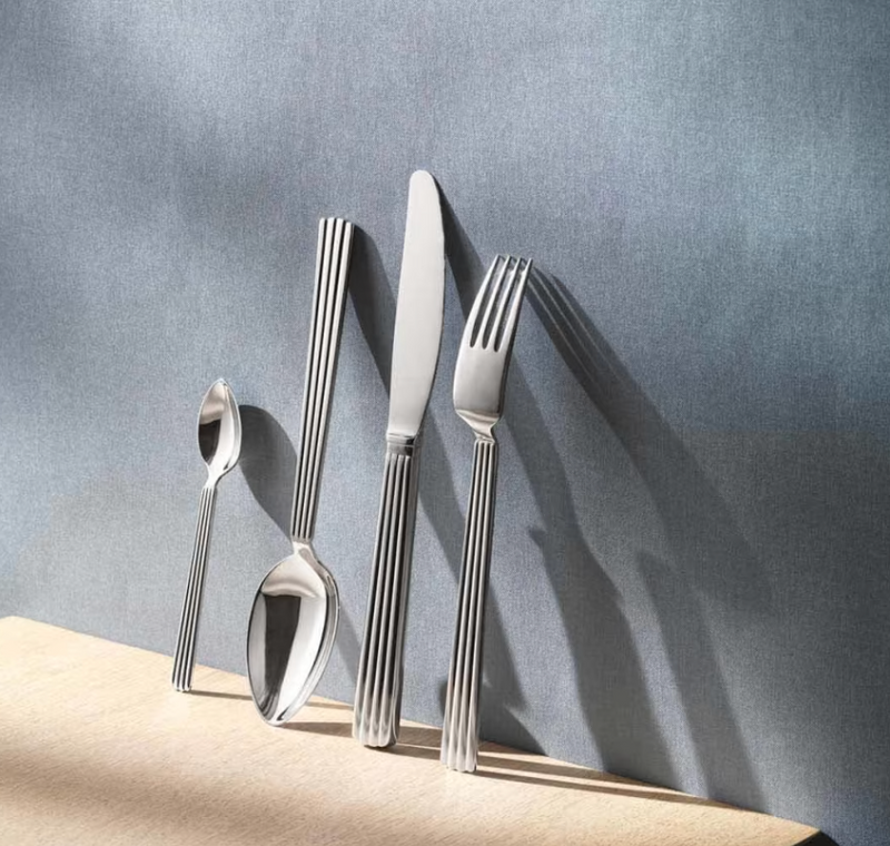 A Georg Jensen Bernadotte Flatware 5 Piece Setting stands upright on a light wooden surface against a textured grey background, each utensil showcasing the signature vertically striped handle design.