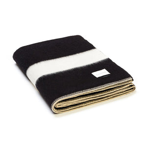 The Siempre Recycled Alpaca Throw by Blacksaw is a heavyweight black and white blanket with a wide white stripe, stitched edges, and a small corner label—perfect as a cozy fireside companion.