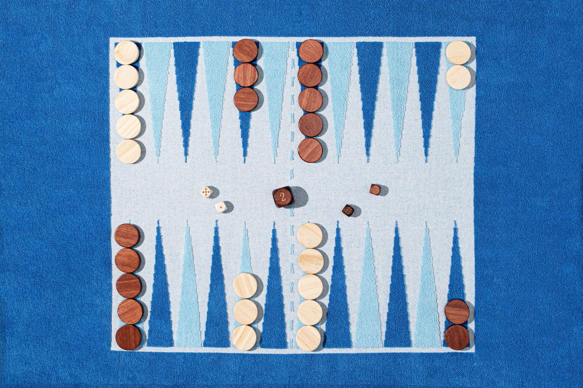 The G. McGee Southampton Beach Backgammon set features light and dark wooden pieces, three dice showing 2, 3, and 1, on a blue and white board next to a plush cotton terry luxury beach towel.