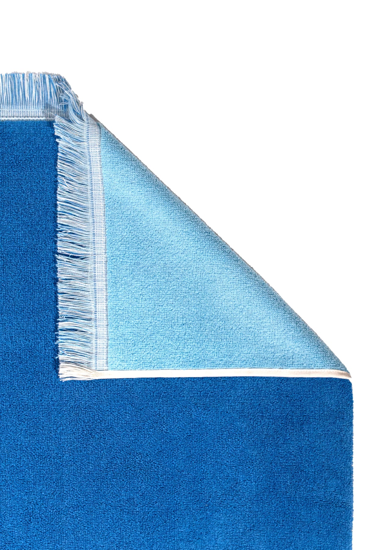 The G. McGee Southampton Beach Backgammon is a blue rectangular luxury beach towel with white trim and fringes on one end. A folded top right corner reveals its plush, lighter blue cotton terry underside, set against a white background.