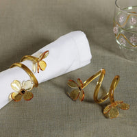 The Botanica Napkin Ring set of 4 by Zodax features gold and enamel butterfly designs displayed on a beige tablecloth, with one ring elegantly holding a white napkin, while a glass adorned with pink flowers in the background enhances this sophisticated botanical table decor.